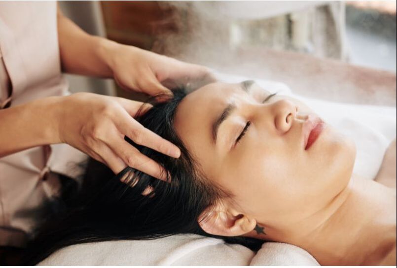 A woman with black hair lays on her back and receives a scalp massage from a massage therapist