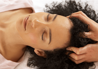 A picture of a woman lying on her back, with curly black hair, receiving a scalp massage