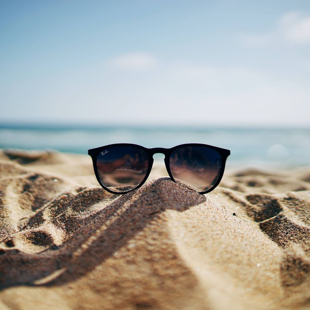 A pair of sunglasses sits on the sand at the beach, with the ocean in the background